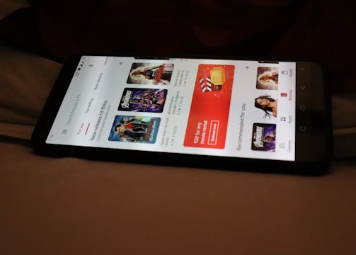 A smartphone displaying a streaming app interface, showing various movie and TV show thumbnails. The screen includes categories like 'For you' and 'New release hit films', with images from popular films. The background is a dimly lit setting with a fabric surface.