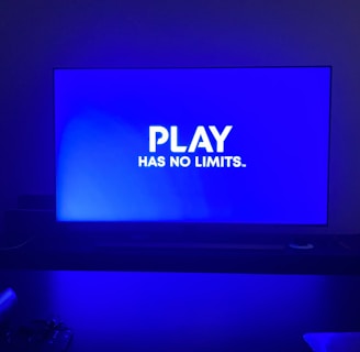 A television screen displays the phrase 'PLAY HAS NO LIMITS.' in bold white text against a blue background. The room is dimly lit with the screen being the primary light source, casting a blue hue on the surroundings. A table in front of the TV has indistinguishable objects that are partially visible in the foreground.