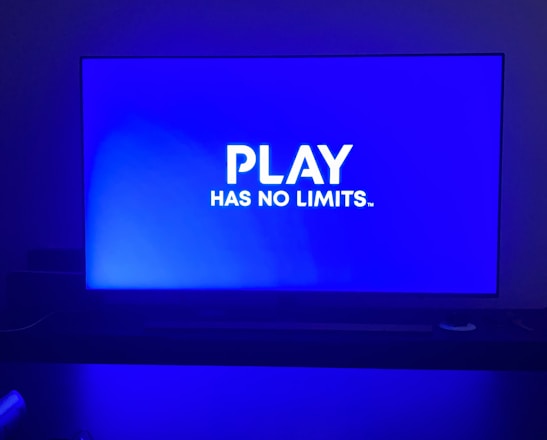 A television screen displays the phrase 'PLAY HAS NO LIMITS.' in bold white text against a blue background. The room is dimly lit with the screen being the primary light source, casting a blue hue on the surroundings. A table in front of the TV has indistinguishable objects that are partially visible in the foreground.