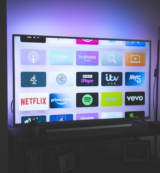 A flat-screen television is displayed in a dimly lit room, showcasing a variety of streaming service apps on its screen, such as Netflix, Prime Video, and Spotify. The TV is positioned on a black stand beside a tall floor lamp, casting a soft ambient light on the surrounding area.