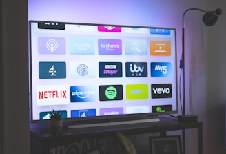 A flat-screen television is displaying a variety of streaming app icons, including Netflix, Prime Video, Spotify, and ITV Hub among others. The TV is placed on a media console with a small potted plant beside it. A tall lamp stands to the right, and a faint, ambient light casts a purple hue on the wall above the TV.