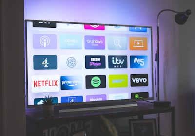 A flat-screen television is displaying a variety of streaming app icons, including Netflix, Prime Video, Spotify, and ITV Hub among others. The TV is placed on a media console with a small potted plant beside it. A tall lamp stands to the right, and a faint, ambient light casts a purple hue on the wall above the TV.