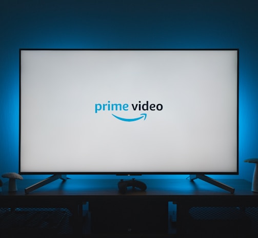 A television screen displays the Prime Video logo, situated on an entertainment unit in a dimly lit room. The room is illuminated with a subtle blue backlight, creating a calming atmosphere. Two small decorative items, resembling mushrooms, are placed on either side of the unit.