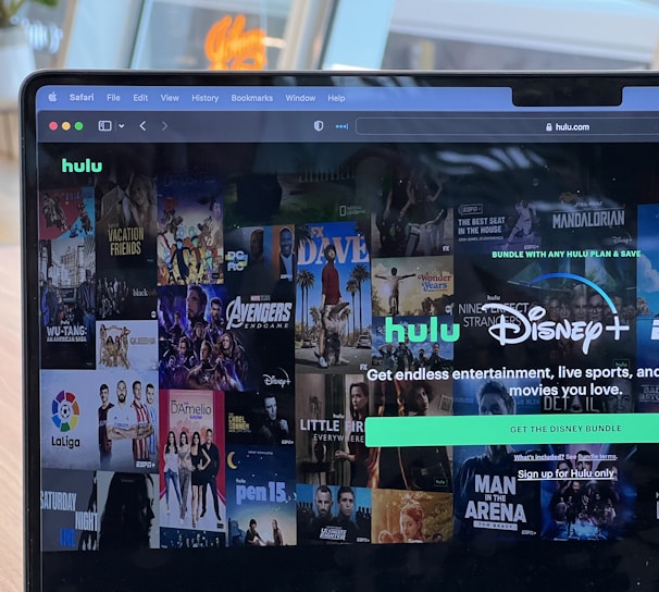 A laptop screen displays a web page featuring a mix of movie and TV show covers, highlighting popular titles and a Hulu and Disney+ bundle promotion. There are visible icons and images of films and shows alongside promotional text offering entertainment and live sports.