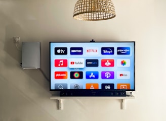 A modern living room features a wall-mounted flat-screen television displaying multiple streaming app icons, such as Apple TV, HBO Max, Netflix, Disney+, and Prime Video. Below the TV is a soundbar sitting on a small white shelf. A wicker pendant light hangs from the ceiling above, and the wall is painted a neutral color.