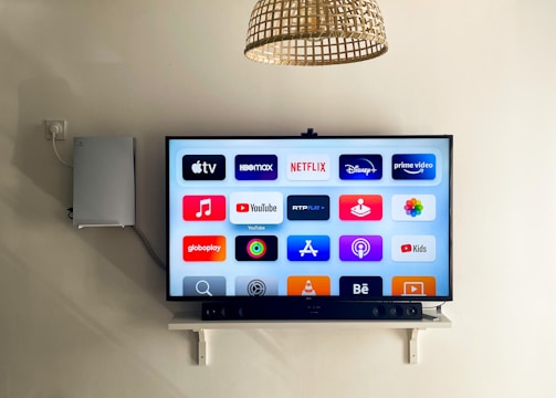A modern living room features a wall-mounted flat-screen television displaying multiple streaming app icons, such as Apple TV, HBO Max, Netflix, Disney+, and Prime Video. Below the TV is a soundbar sitting on a small white shelf. A wicker pendant light hangs from the ceiling above, and the wall is painted a neutral color.