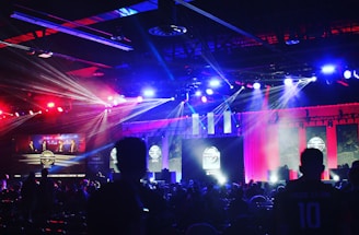A dimly lit indoor arena filled with an audience. Bright stage lights in red and blue illuminate multiple large screens displaying graphics.