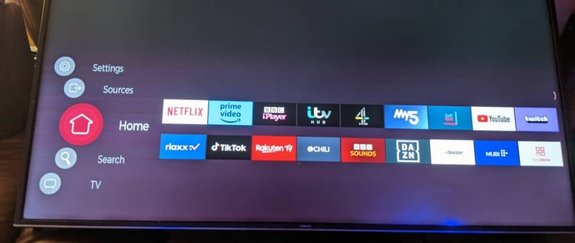 A television screen displays a smart TV interface with multiple app icons arranged in rows. Options such as Settings, Sources, Home, Search, and TV are listed on the left side. Some recognizable app icons include Netflix, Prime Video, BBC iPlayer, ITV Hub, All 4, My5, YouTube, and TikTok. The time and date are shown in the top right corner.
