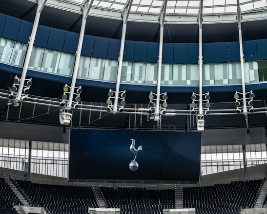 A modern stadium interior featuring a large digital screen with a logo prominently displayed. The seating areas are empty, with dark-colored seats and some structural elements like beams and glass walls visible. A combination of dark blue and neutral tones are prominent.