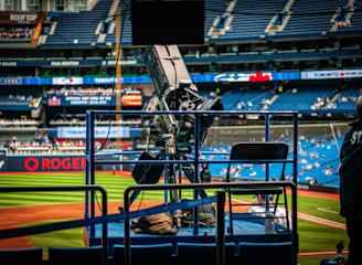 A professional-grade video camera is mounted on a platform overlooking a baseball field. The stadium seats in the background are filled with some spectators, and various advertisements are visible around the field. A staff member wearing a jacket with 'STAFF' written on the back is positioned near the camera.