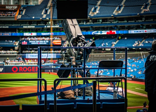 A professional-grade video camera is mounted on a platform overlooking a baseball field. The stadium seats in the background are filled with some spectators, and various advertisements are visible around the field. A staff member wearing a jacket with 'STAFF' written on the back is positioned near the camera.