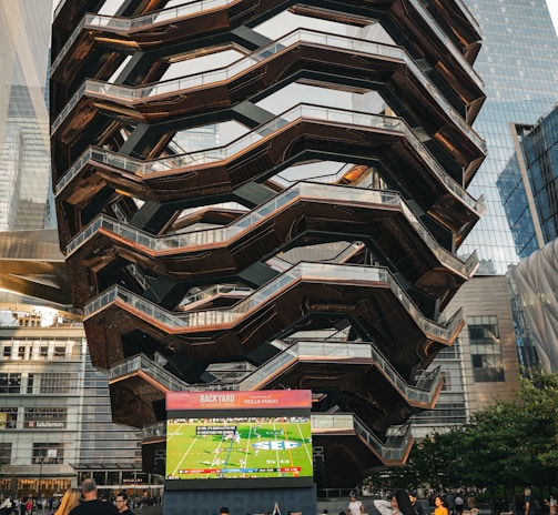 A large, spiral architectural structure with multiple levels stands prominently in an urban setting. In front of the structure, a large screen displays a sports broadcast, and several people are gathered around, some sitting and others standing. The scene is set outdoors, with towering modern buildings surrounding the area. The ground is paved with a geometric pattern of gray and white tiles.