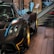 A sleek and luxurious sports car with a carbon fiber body and gold alloy wheels is displayed on a polished wooden platform. The setting includes glass barriers, informational plaques, and a staircase in the background, creating a sophisticated showroom environment.